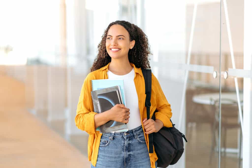 Woman university student. Happy lovely brazilian or hispanic female student, with a backpack, hold books and notebooks in her hand, stand near the university campus, looks and smile to the side