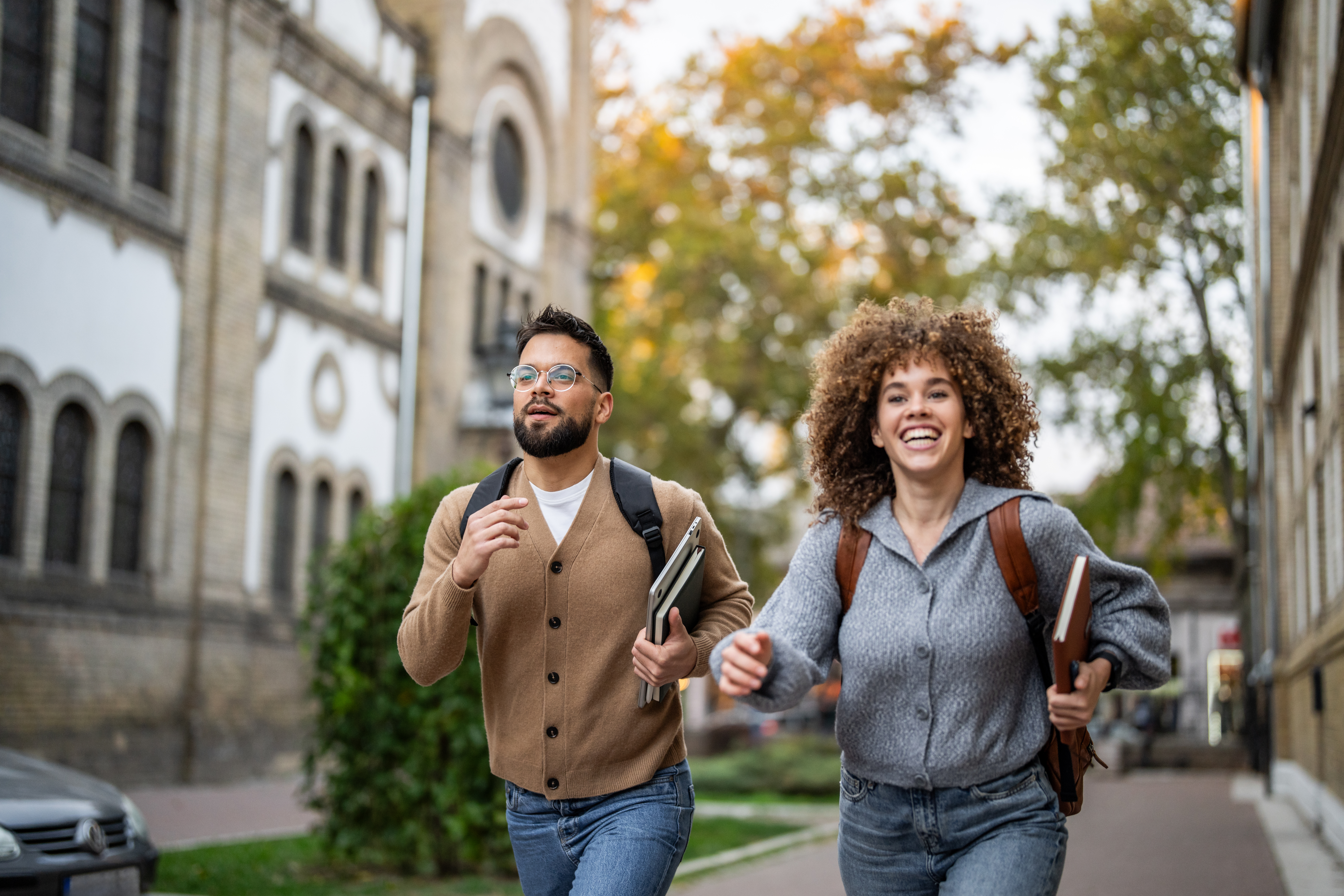 Two cheerful multi-ethnic students running in the university campus, holding books and backpacks, probably late for class, smiling and looking forward