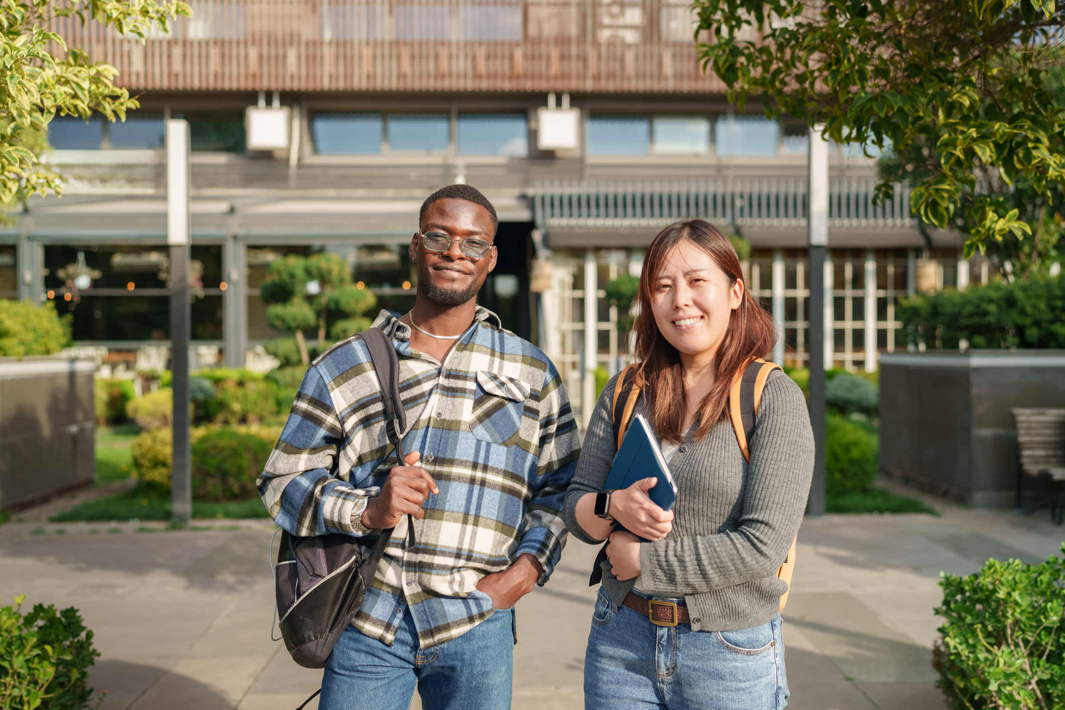 Portrait of two diverse college students smiling confidently outdoors on campus, carrying backpacks and study materials. Represents global education, youth and friendship.