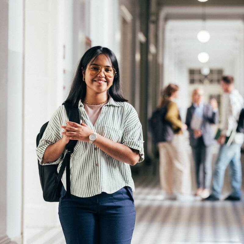 A confident student with a backpack and glasses walks through a bright university hallway. In the background, classmates are engaged in discussion, highlighting a vibrant academic environment.